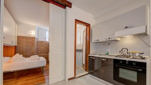 A compact kitchen with a stove, oven, and white cabinets. A bed with white linens is visible in the adjacent room. - Vicolo 42 At 200m From Lake (Salò)