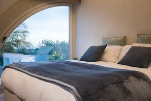 Un dormitorio con techo curvo, cama con ropa de cama blanca y gris oscuro, y vista a zonas verdes al exterior.