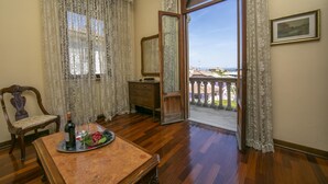 Ein Zimmer mit Holzboden, einem Esstisch mit Obsttablett, einem Holzstuhl, einem Schminktisch und einem großen Fenster mit Vorhängen.
