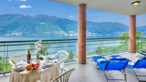 A balcony with a table set for dining, overlooking a mountainous landscape and a body of water. - Va Pensiero sul Lago Stunning View (Tronzano Lago Maggiore)