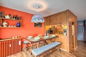 Un coin repas avec une table en bois dressée pour quatre personnes, un banc et un espace cuisine doté de meubles en bois et d’un luminaire suspendu.