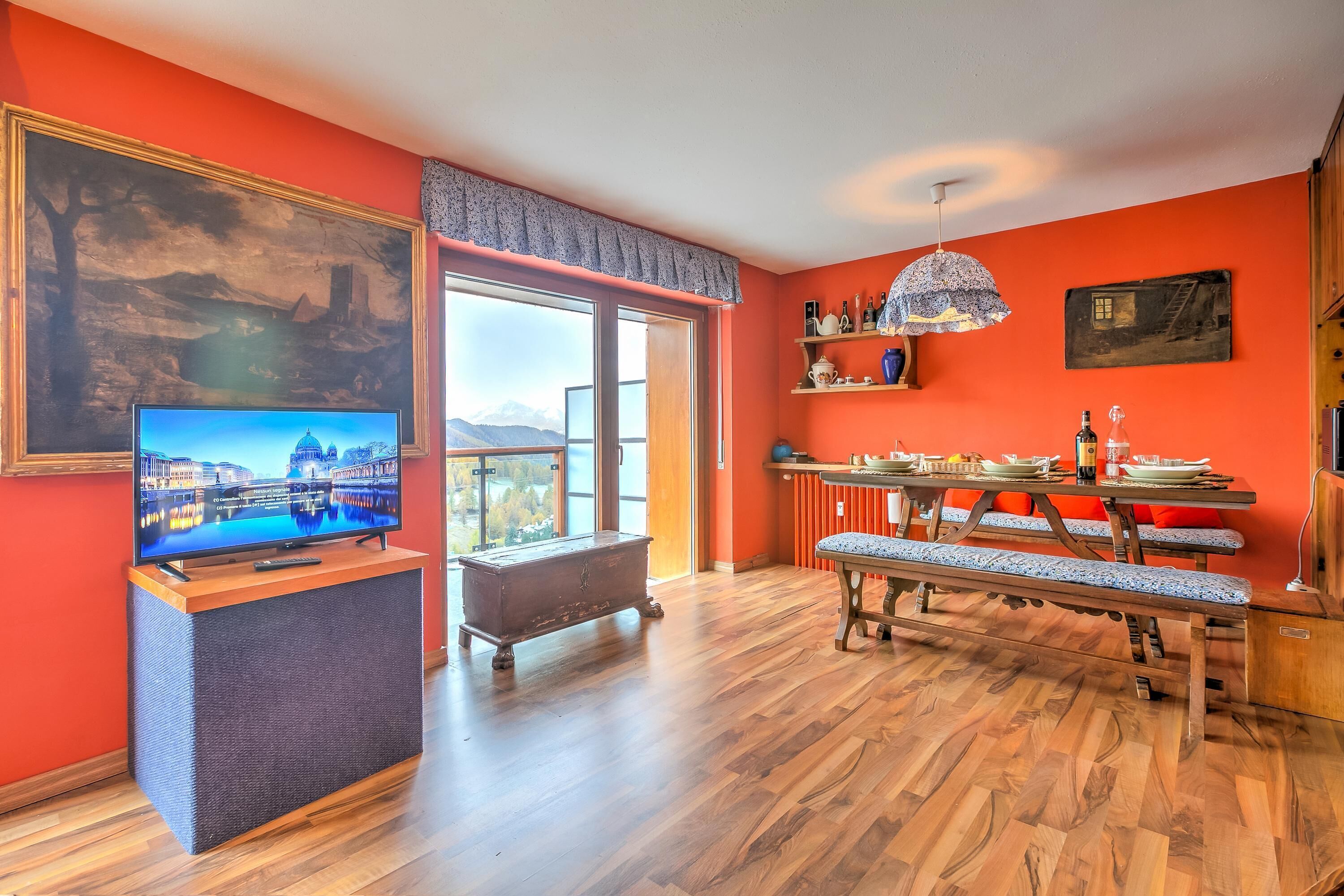 Una habitación con piso de madera, un televisor sobre un mueble, un banco, una mesa de comedor con sillas y un ventanal con vistas a montañas.