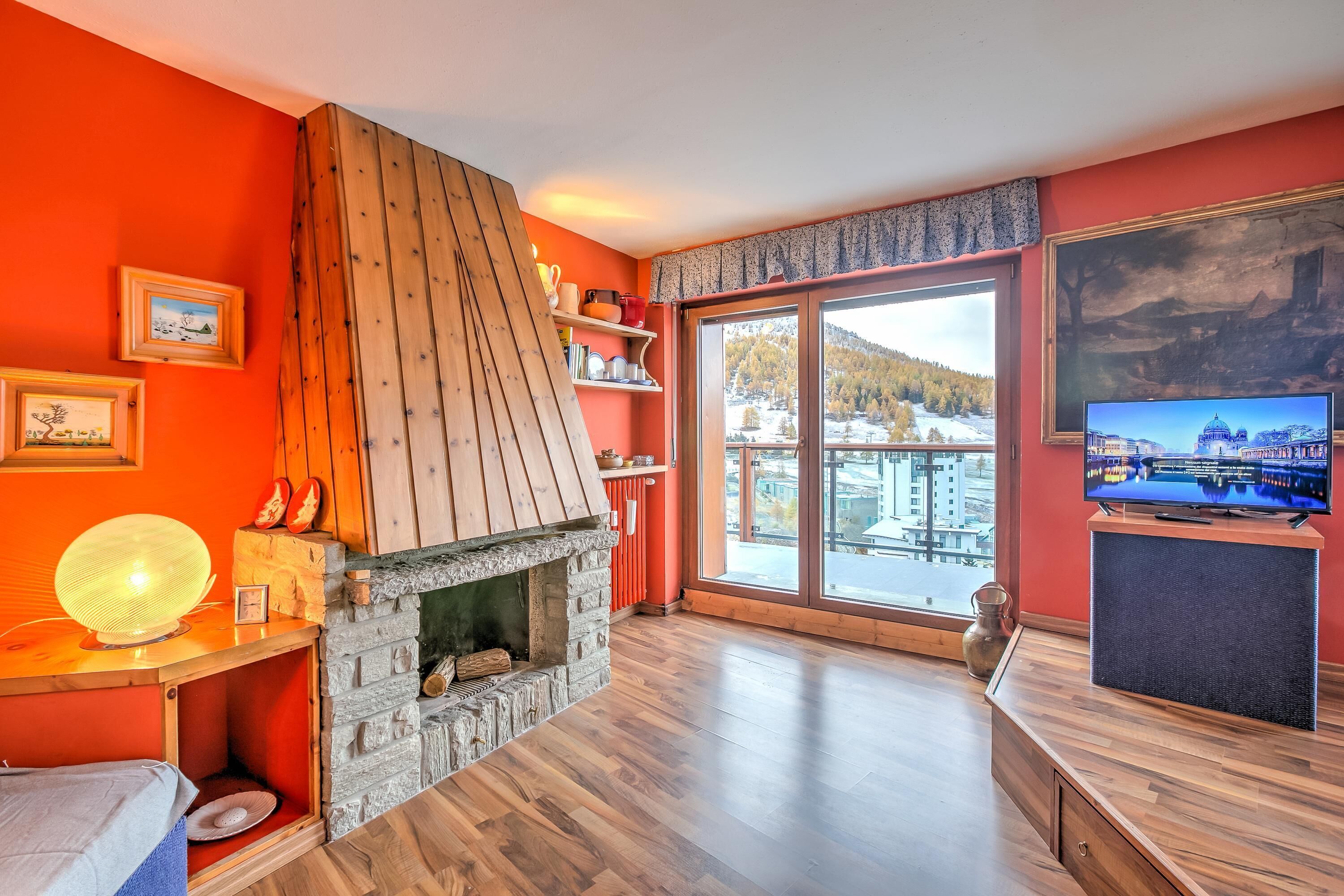 A room with a stone fireplace, wooden shelves, a large window with a view, and a television on a wooden stand.