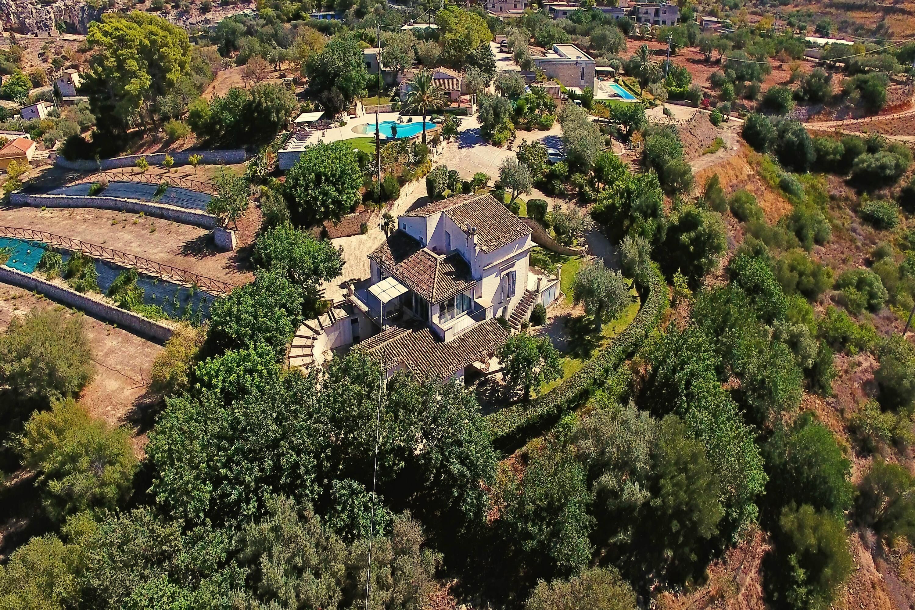 An aerial view of a residential area with a large house featuring a tiled roof, a swimming pool, and surrounding greenery.
