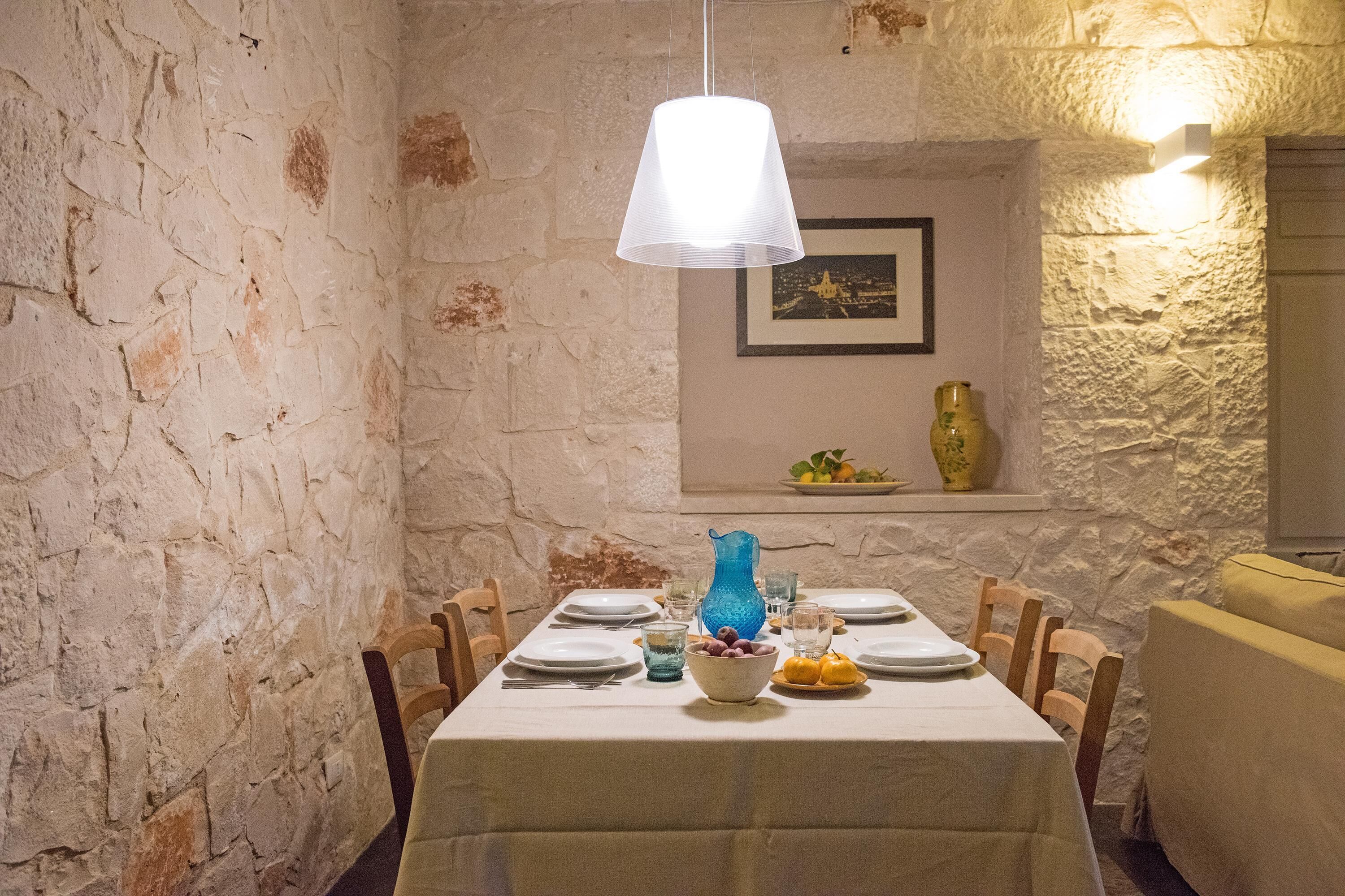 Un coin repas avec un mur en pierre, une nappe blanche et un vase bleu.