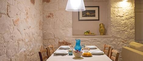 Un coin repas avec un mur en pierre, une nappe blanche et un vase bleu.