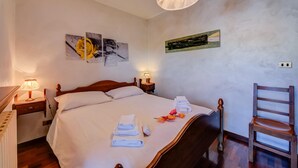 A neatly made bed with towels and flowers, two bedside lamps, a wooden chair, and artwork on the walls. - Sestriere Terrace Apt Few Steps From Ski (Sestriere)