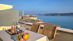A table with a bottle of olive oil, glasses, and a fruit bowl on a balcony overlooking a coastal town with a prominent tower. - Residenza Faust-historical Center Sal (Salò)