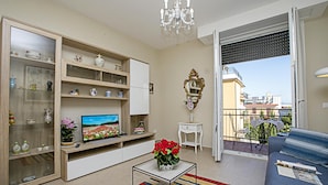 Sala de estar con televisor, sofá, mesa de centro con flores, estantería y balcón con vistas.