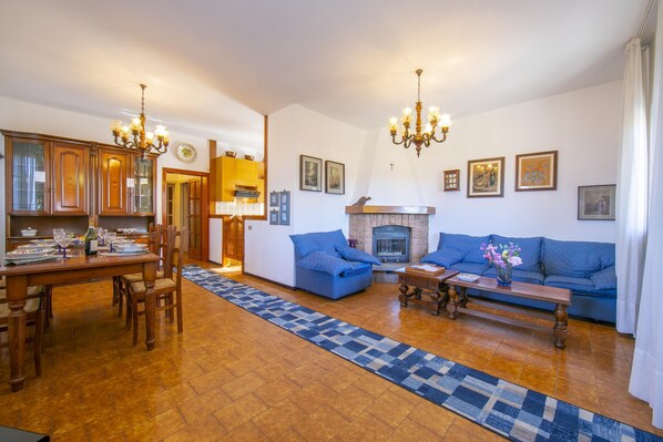 A spacious living room with a fireplace, blue sofas, a dining area with wooden furniture, and a patterned rug.