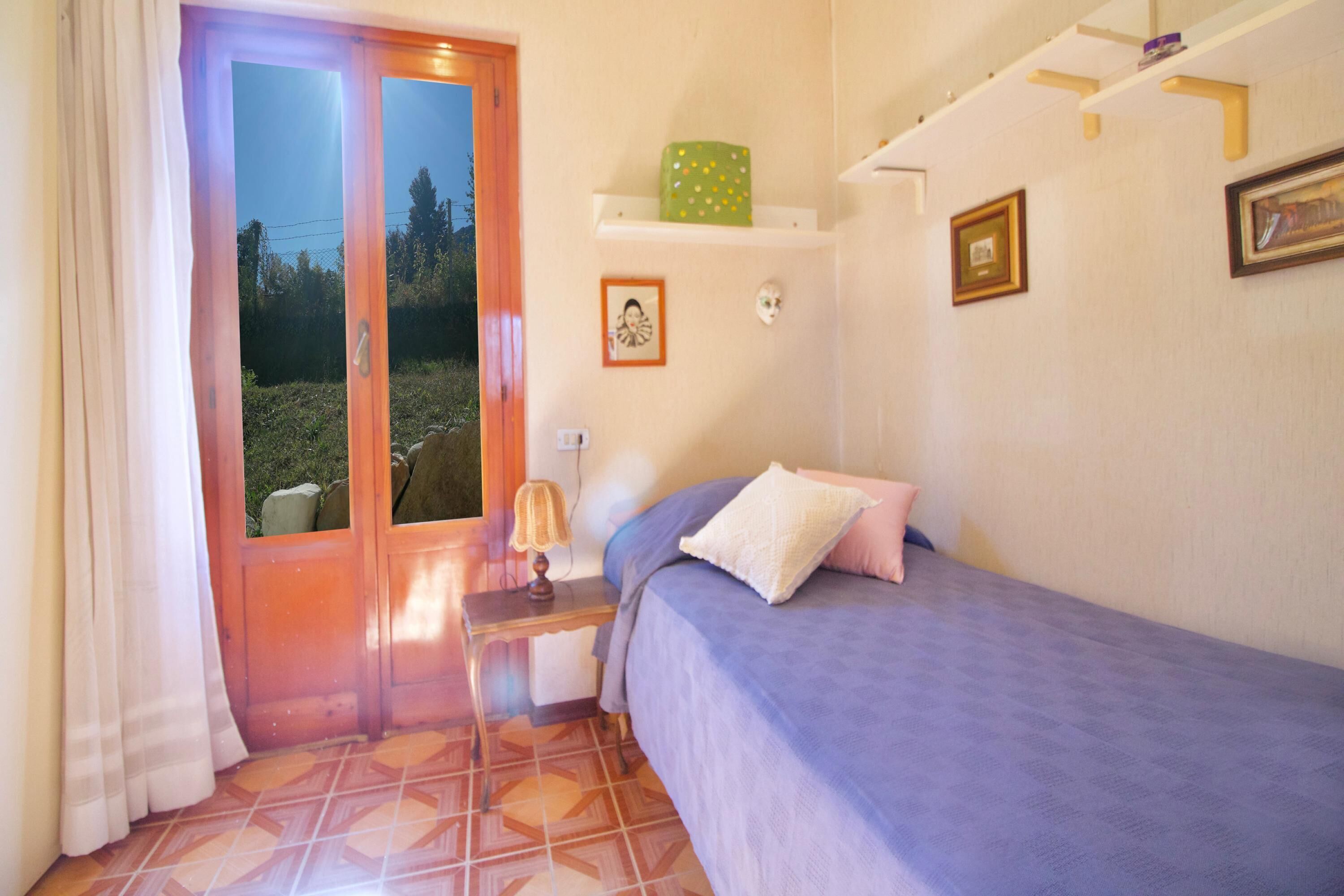A bedroom with a bed, a nightstand, a lamp, a window with curtains, and a wall with a shelf and framed pictures.