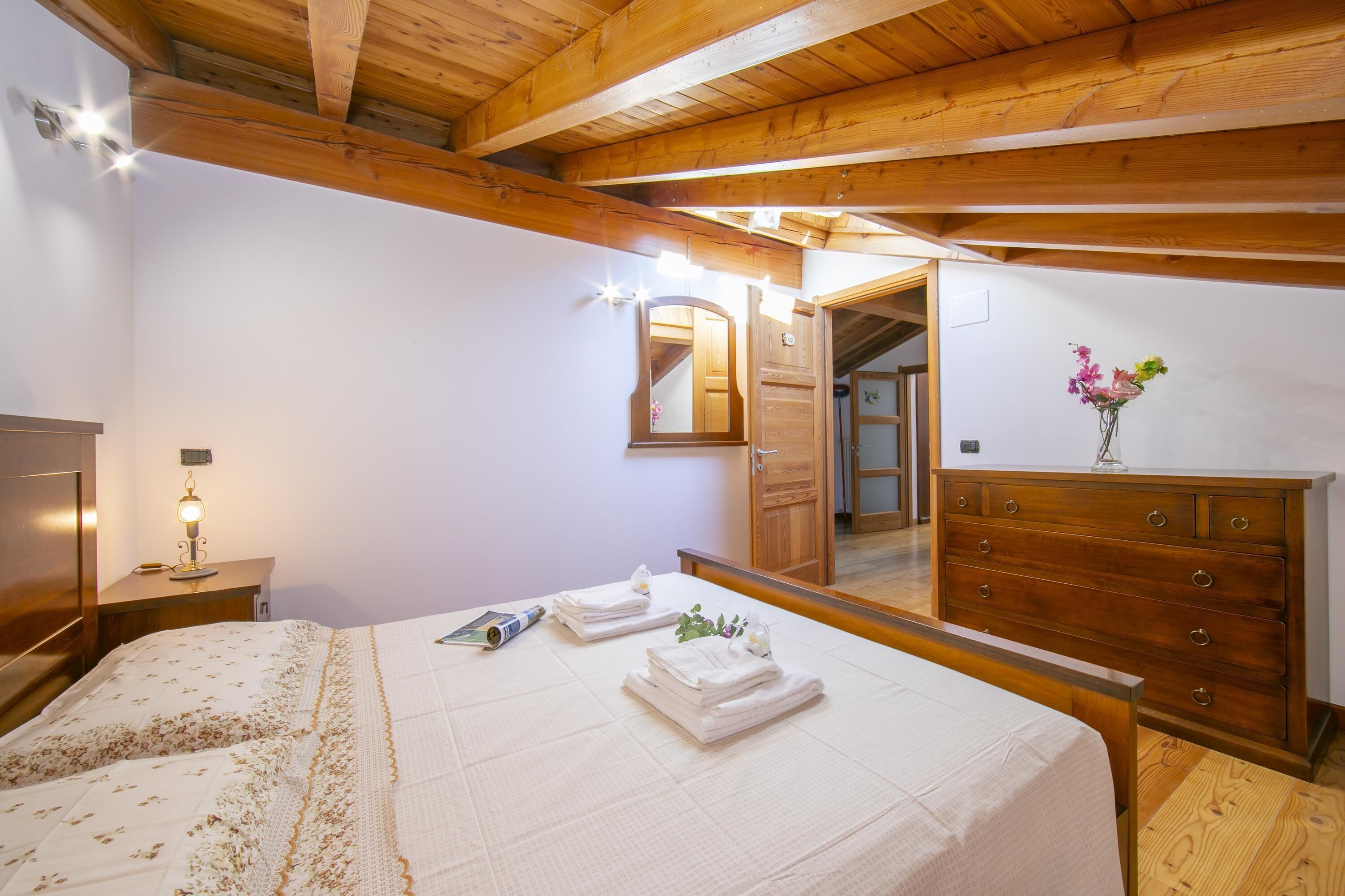Una camera da letto con soffitto in legno, un letto con copriletto floreale, un comò con specchio e un vaso con fiori sul comò.
