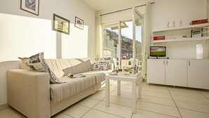 A living room with a sofa, a coffee table, and a TV on a stand. There are framed pictures on the wall and a window with a view of a hill. - Torbole Relax Pool Balcony Apartment (Nago-Torbole)