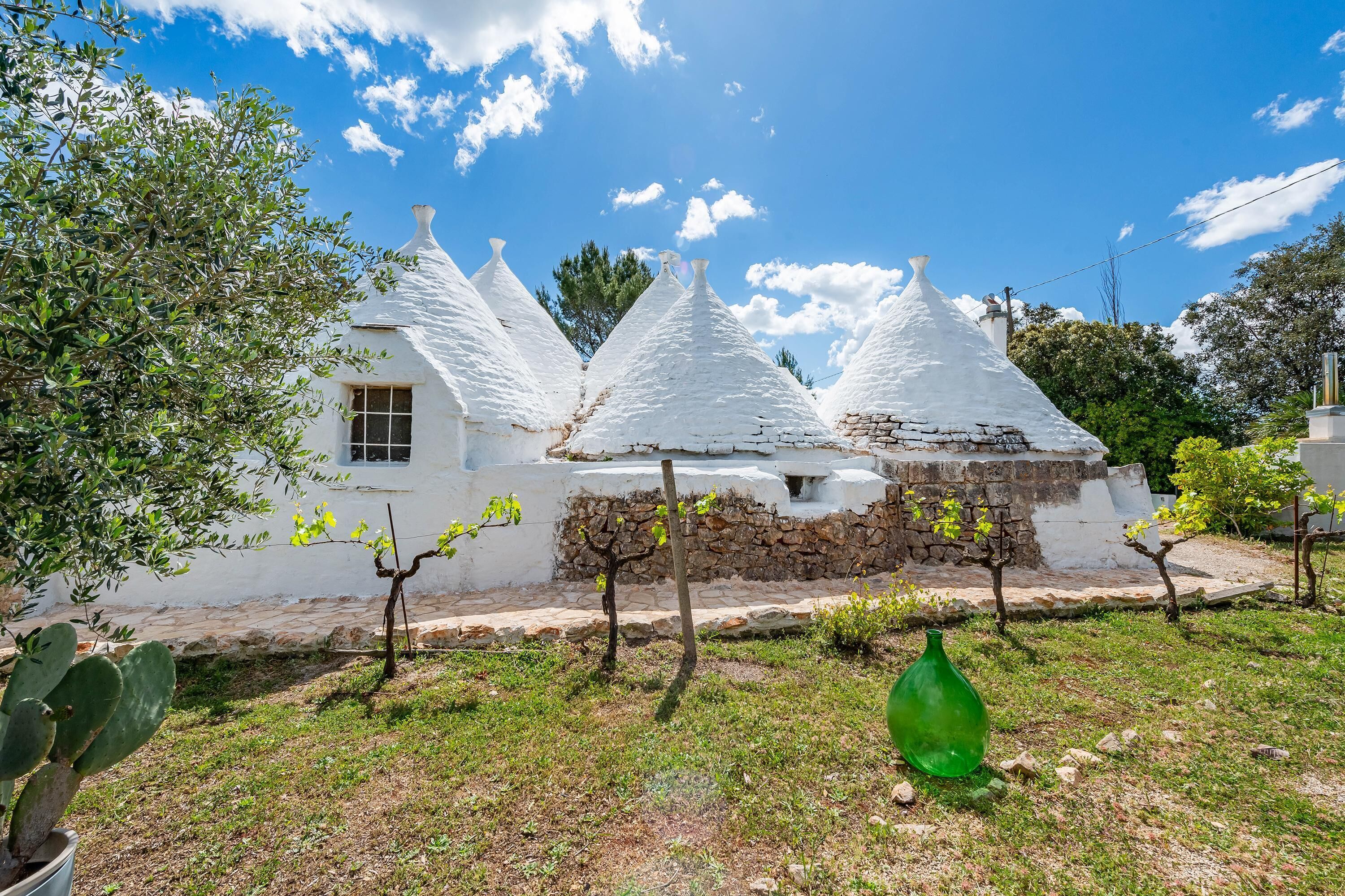 A white building with multiple conical roofs, a stone wall, and a cactus plant in the foreground.