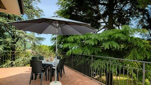 Een terras buiten met een tafel, stoelen, een grote parasol en een metalen hekwerk.