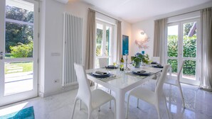 A dining area with a glass table, white chairs, and a view of greenery outside. - Villa Victor 100m From sea (Massa)