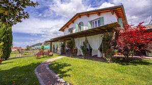 A two-story house with a covered porch, a brick pathway leading to the entrance, and a well-maintained garden with various plants and flowers. - Villa Clara few km From the sea (Pietrasanta)