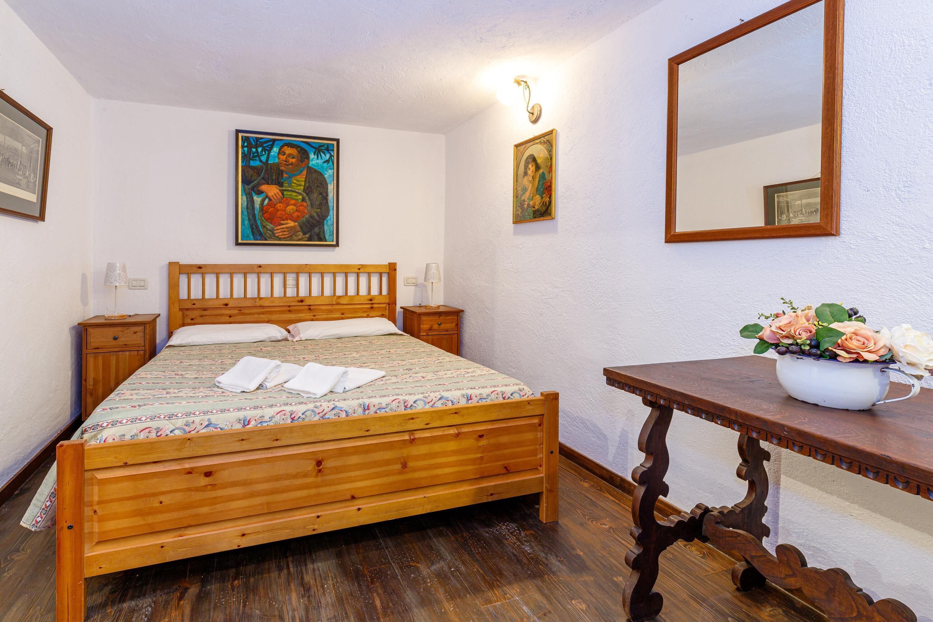 A bedroom with a wooden bed, a nightstand, a bedside table with a vase of flowers, and two wall-mounted paintings.