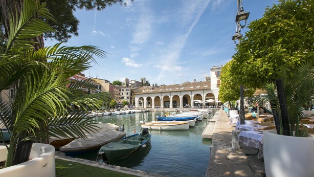 A canal with boats docked, a building with arches, and potted plants along the water's edge. - Villa Mimosa - Appartamento 1 (Desenzano del Garda)