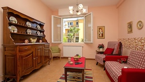 A living room with a wooden cabinet, a red sofa, a table with a red tablecloth, and a window with a view of buildings. - Villa Mimosa - Appartamento 1 (Desenzano del Garda)