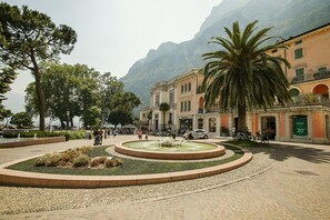 Uma praça pública com uma fonte circular, rodeada por árvores e edifícios com varandas.