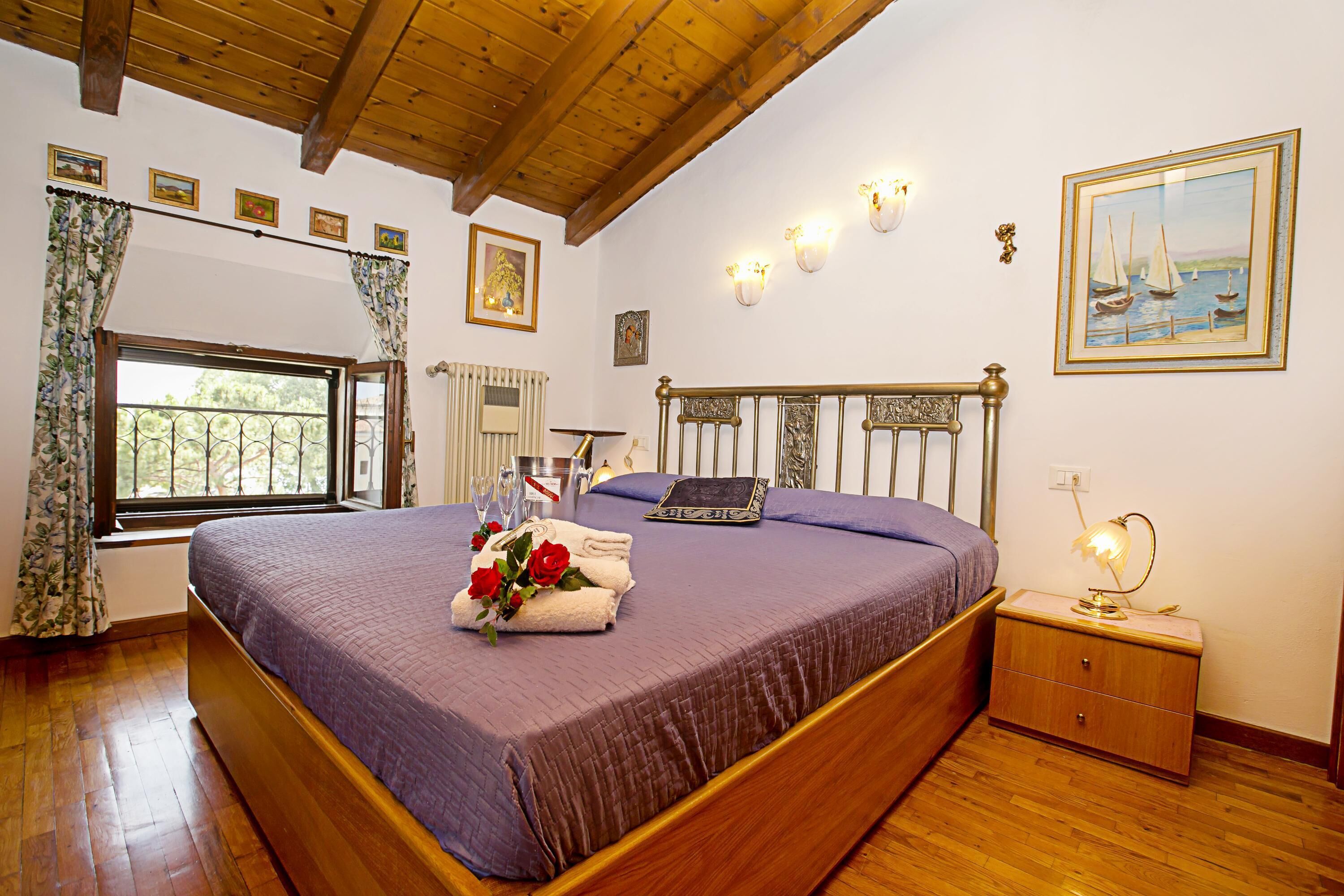 A bedroom with a wooden ceiling, a large bed with a purple bedspread, a nightstand with a lamp, and a window with curtains.