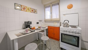 Eine kleine Küche mit weiß gefliesten Wänden, einer weißen Arbeitsplatte, einem orangefarbenen Schrank, einem weißen Herd und einem Fenster mit Spitzendeckchen.