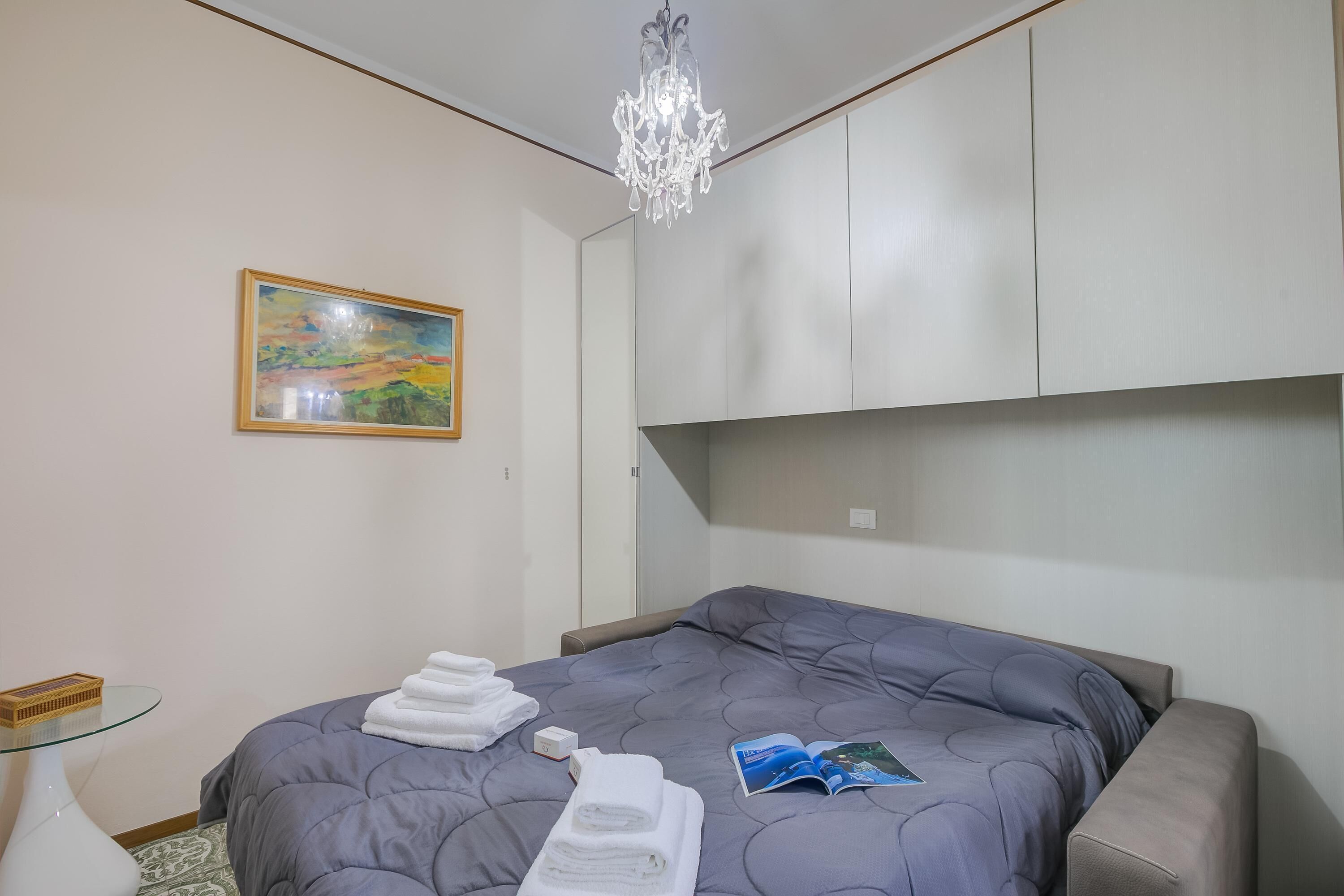 A modern hotel room with a large bed, a chandelier, a painting, and neatly folded towels.