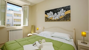 Ein Hotelzimmer mit grüner Bettdecke, einem Blumenbild an der Wand, einem Fenster mit Blick auf Gebäude und einem Nachttisch mit Lampe.