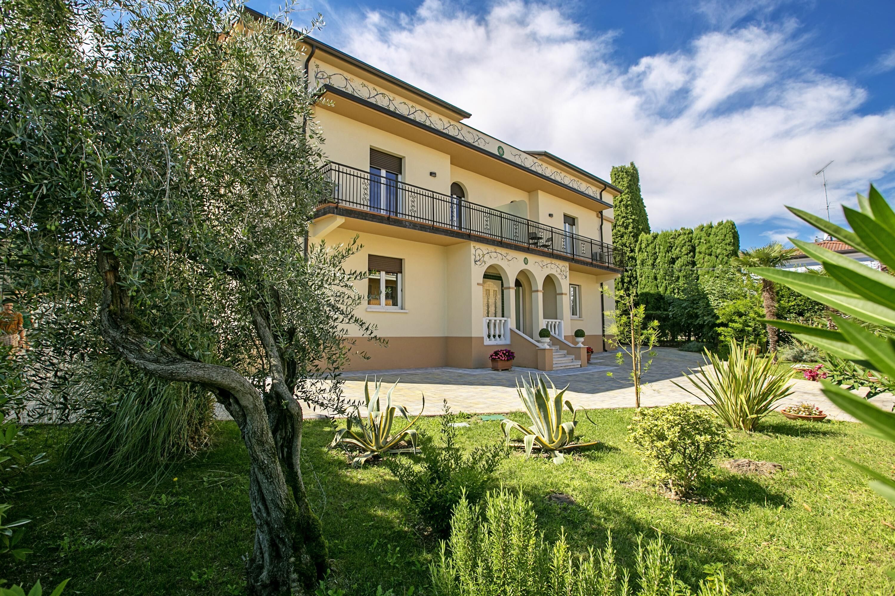 A two-story building with a balcony, arched windows, and a garden with various plants.