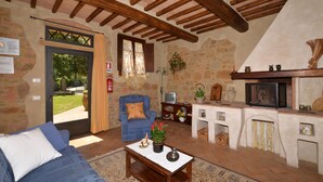 A cozy living room with a stone wall, wooden beams, a blue sofa, a coffee table with decorative items, a fireplace, and a television. - Villa Casacheta in Montaione (Montaione)