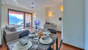 A dining area with a table set for four, a sofa with cushions, a TV in the background, and a balcony with a view. - Panorama Fantastico Apt Pool Beach (Ghiffa)