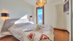 A bedroom with a bed, a book, towels, a blanket with a heart design, a chandelier, and a window with a view. - Panorama Fantastico Apt Pool Beach (Ghiffa)