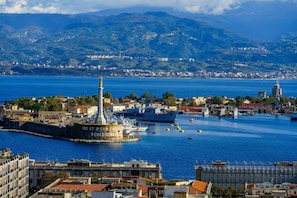 Un paisaje urbano costero con un faro destacado, un puerto con barcos y montañas al fondo.