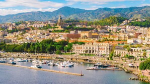 Un paisaje urbano costero con un puerto deportivo, edificios históricos y una cadena montañosa al fondo.