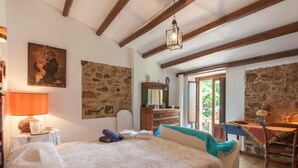 Un dormitorio con cama, cómoda, espejo y escritorio con silla. Se ve follaje verde a través de la ventana.