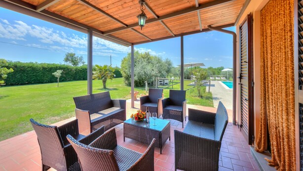 A covered patio with wicker furniture, a table with a vase and glasses, and a view of a swimming pool and greenery.