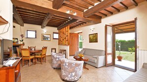 A spacious living room with a high ceiling, wooden beams, a sofa, two armchairs, a dining table with chairs, and a sliding glass door leading to a garden.