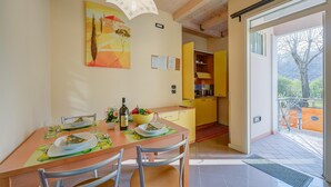 A dining area with a wooden table set for two, a yellow kitchen in the background, and a view of the outdoors through a glass door. - Yellow Apartment Desenzano With Smart TV (Desenzano del Garda)