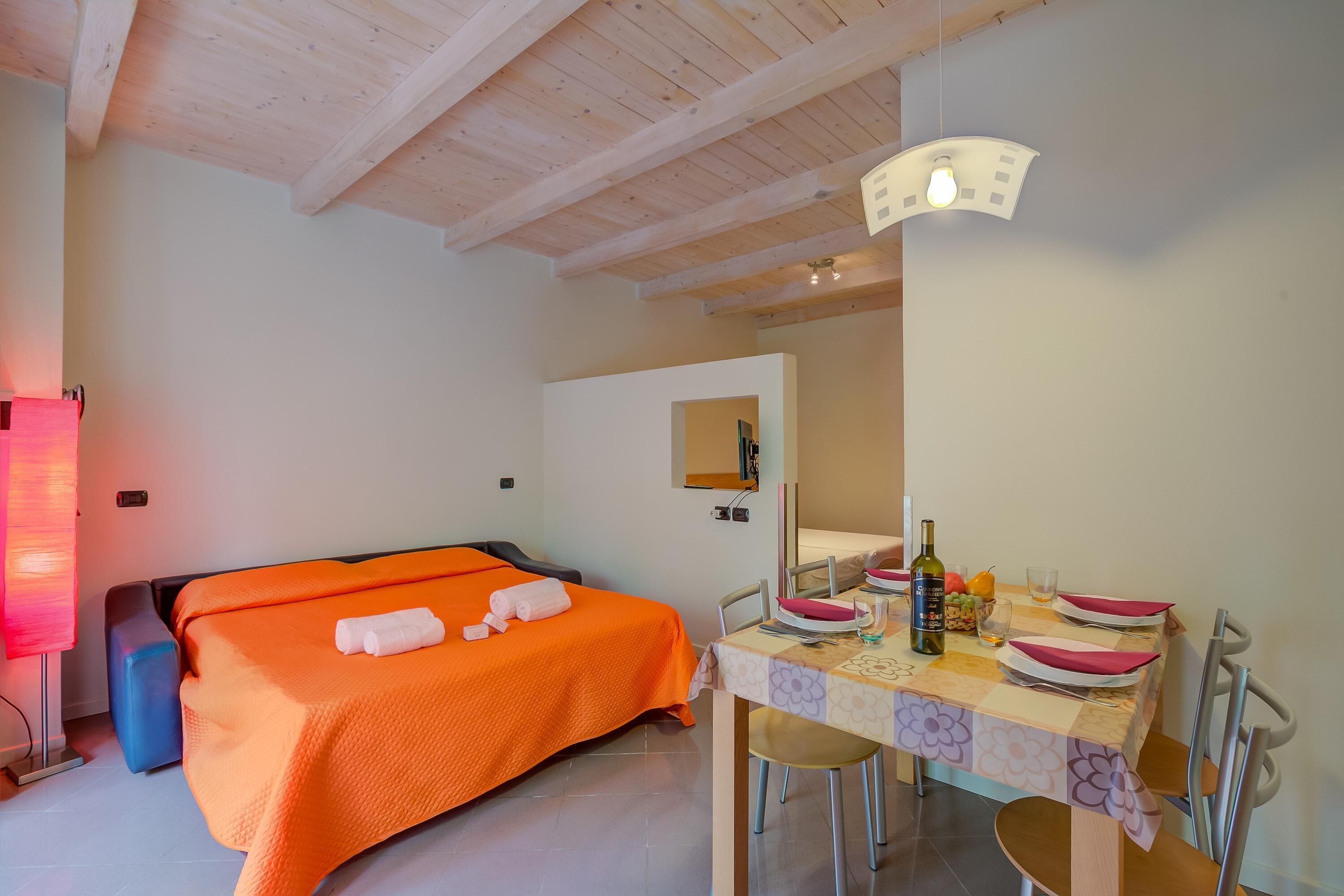 Ein Schlafzimmer mit orangefarbener Bettdecke, einem Nachttisch mit Flasche und Gläsern sowie einem Essbereich mit gedecktem Tisch für zwei Personen.