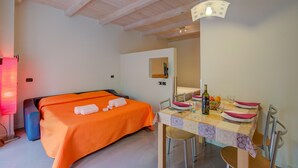 Une chambre à coucher avec une couvre-lit orange, une table de chevet sur laquelle se trouvent une bouteille et des verres, et un coin repas avec une table mise pour deux personnes.