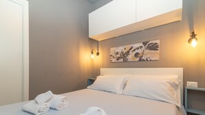 Een keurig opgemaakt bed met opgerolde handdoeken, een hoofdbord en een aan de muur bevestigd kunstwerk.