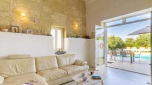 A modern living room with a beige leather sofa, a white coffee table, and a glass door leading to an outdoor pool area. - Ta Patee Pool Farmhouse in Gozo (San Lawrenz)