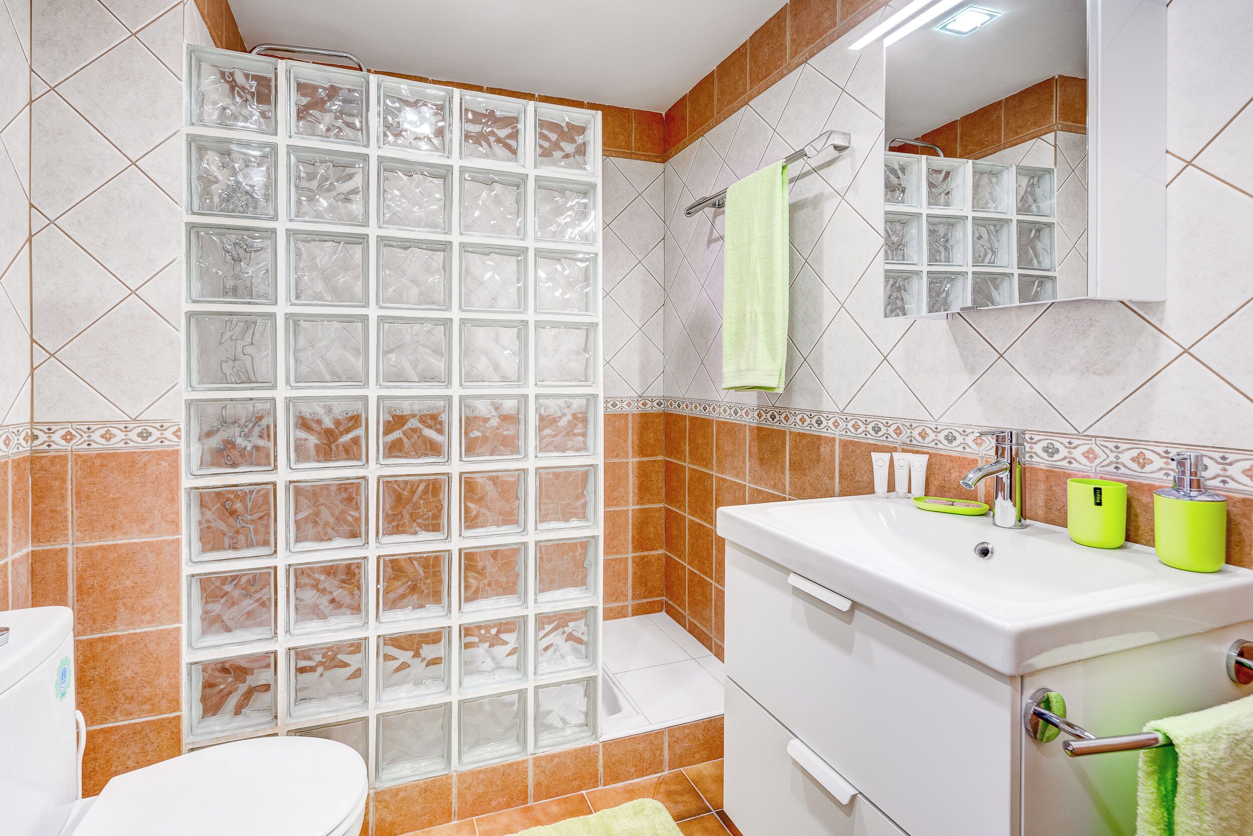 Un baño con cabina de ducha de vidrio, un lavamanos blanco con dispensadores de jabón verdes y un inodoro blanco.