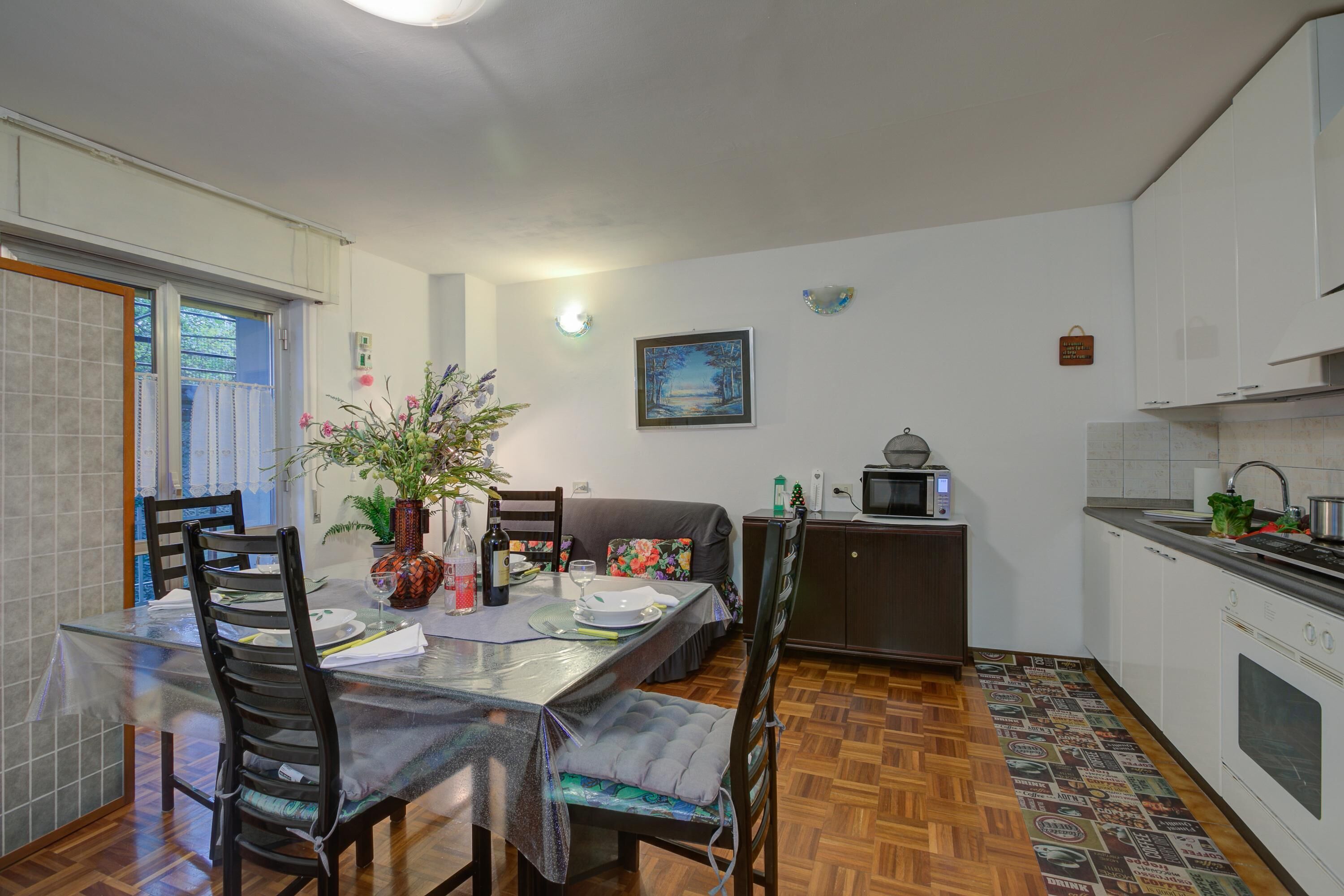Un comedor con una mesa de vidrio, sillas y una planta en maceta. Se ve un sofá y un mueble bajo al fondo.