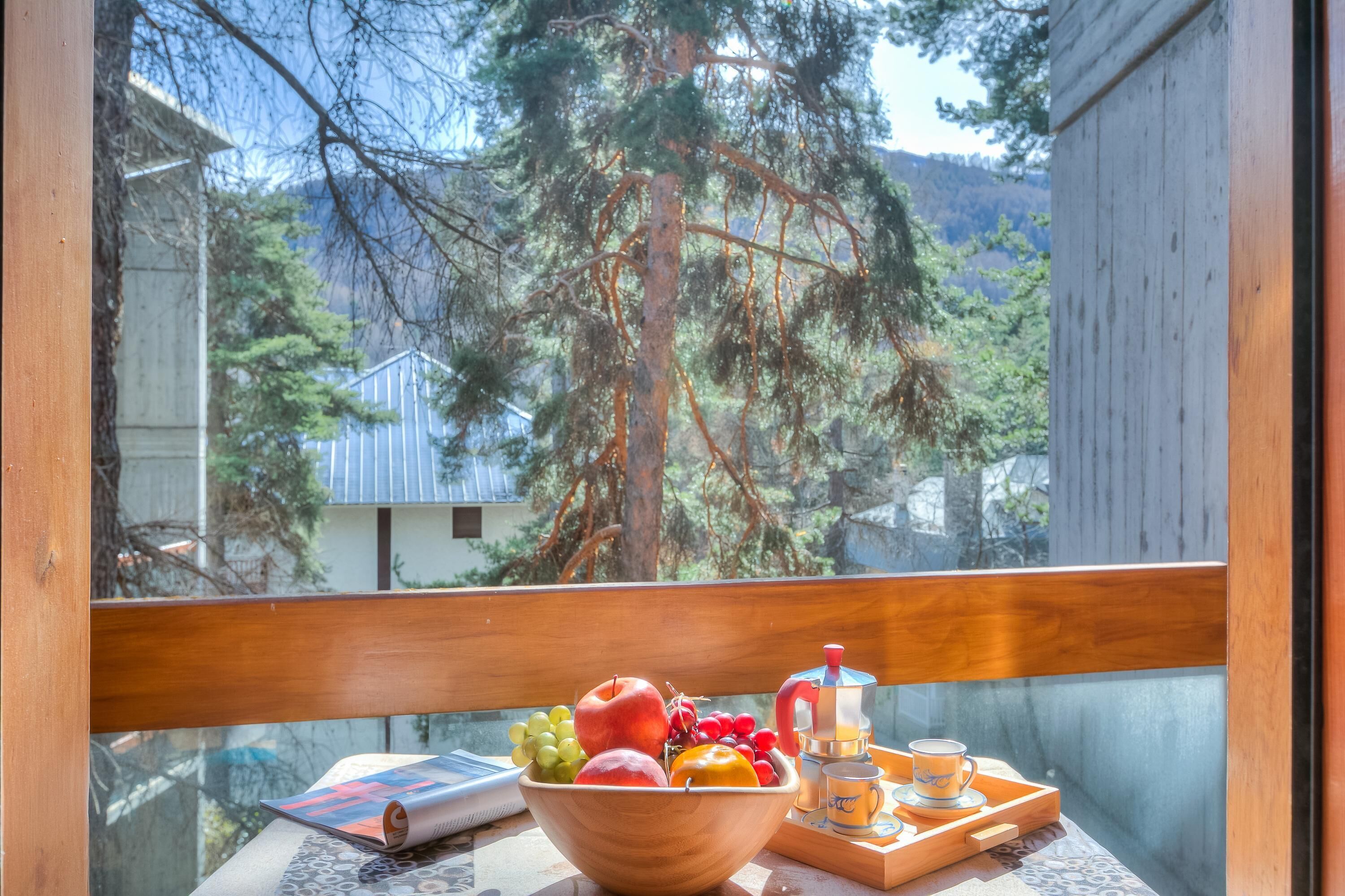 Ein Holztisch mit Obstschale, einem Buch, einer Kaffeemaschine und einer Servierplatte mit Tassen und Untertassen auf einem Balkon mit Blick auf Bäume und Häuser.