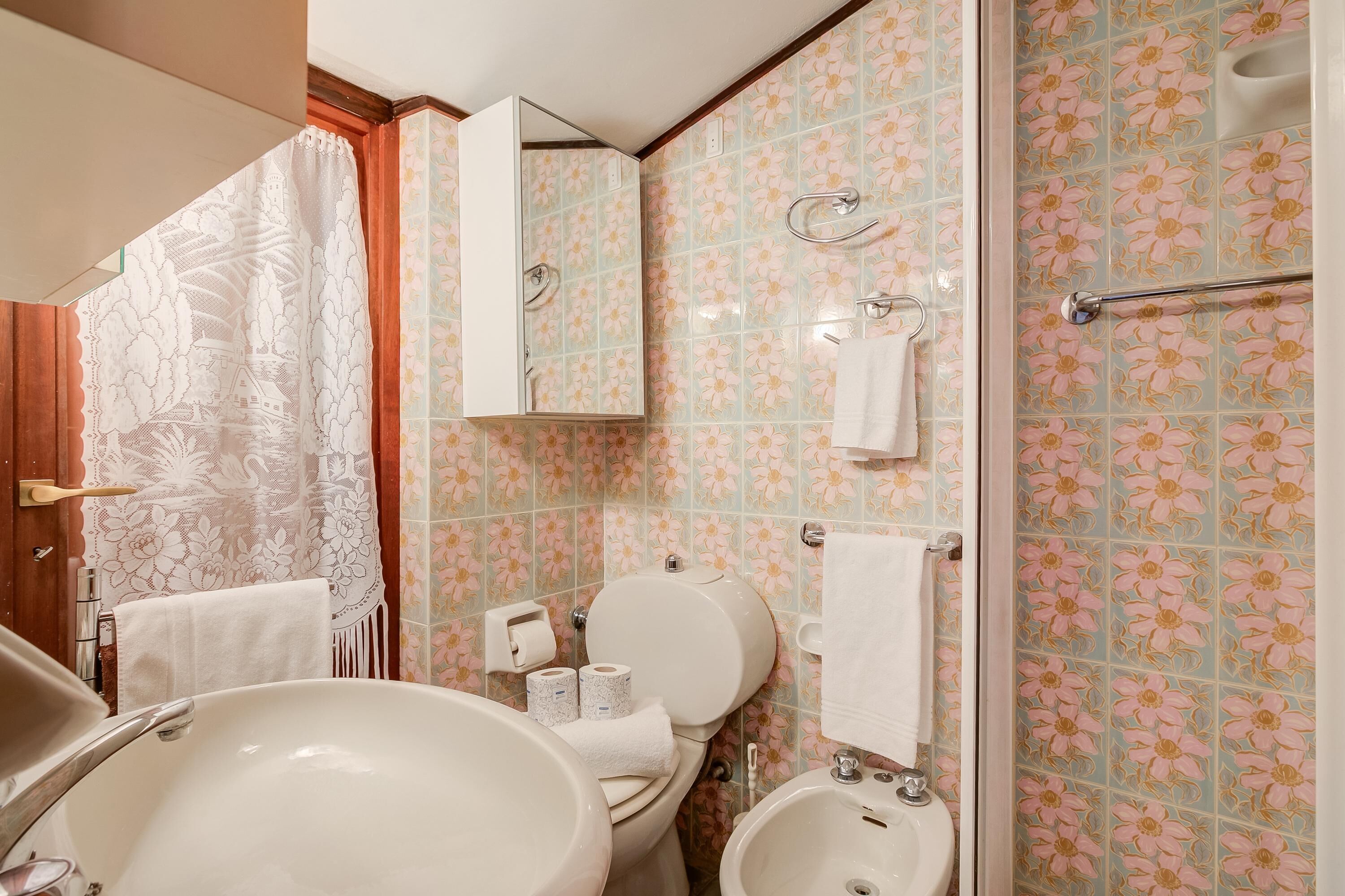 A bathroom with a large sink, toilet, and bidet. It has floral-patterned tiles and a mirror above the toilet.