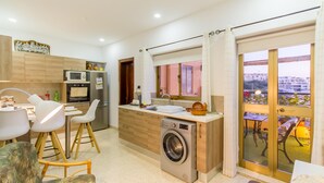 A modern kitchen with a built-in washing machine, dining area with chairs, and a view through a glass door. - Madonnina Sunny Townhouse in Kalkara (Kalkara)