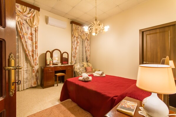 A hotel room with a bed, a desk, a chair, a lamp, and a door.