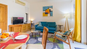 A living room with a sofa, a dining table with a bowl of fruit, a television, and a lamp. - The Strand - Apartment By The Sea Sliema (Sliema)